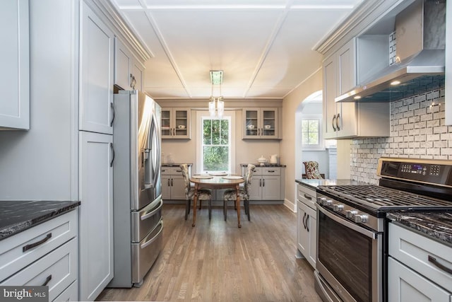 kitchen with appliances with stainless steel finishes, dark stone counters, hanging light fixtures, light hardwood / wood-style floors, and wall chimney range hood