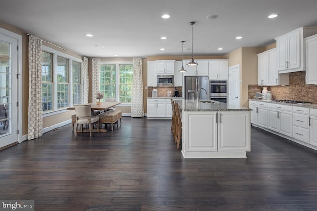 kitchen featuring stone counters, white cabinetry, stainless steel appliances, a center island with sink, and decorative light fixtures