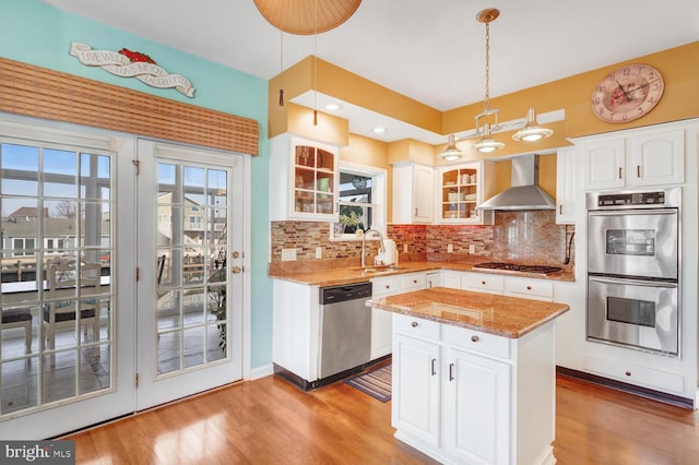 kitchen with wall chimney exhaust hood, sink, hanging light fixtures, stainless steel appliances, and white cabinets