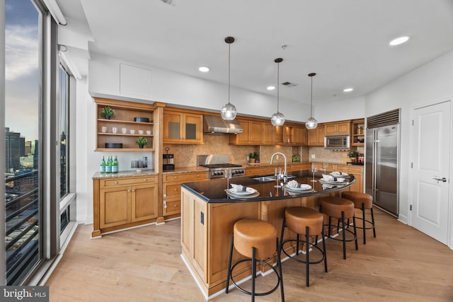 kitchen with sink, built in appliances, an island with sink, decorative light fixtures, and wall chimney exhaust hood