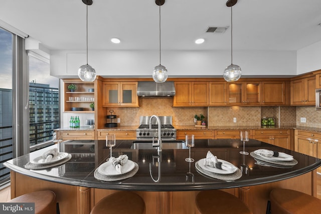 kitchen with wall chimney range hood, decorative light fixtures, stainless steel range, and a center island with sink