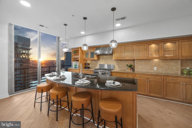 kitchen featuring sink, a center island with sink, hanging light fixtures, wall chimney range hood, and stove