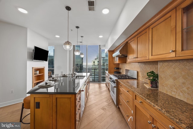 kitchen with sink, tasteful backsplash, an island with sink, stainless steel appliances, and wall chimney range hood