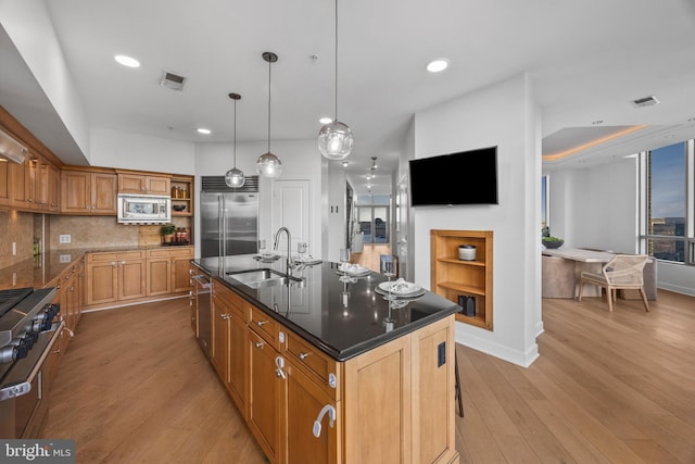 kitchen featuring sink, a kitchen island with sink, hanging light fixtures, built in appliances, and light hardwood / wood-style floors