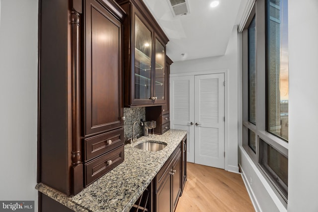 bar with sink, dark brown cabinets, light stone counters, light hardwood / wood-style floors, and decorative backsplash