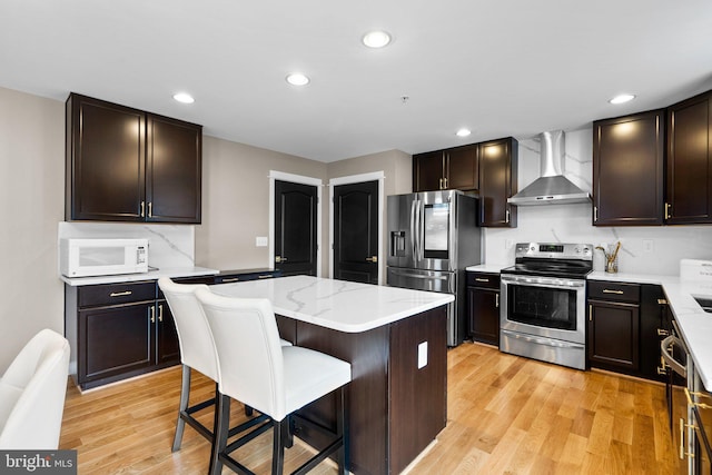 kitchen with wall chimney range hood, a kitchen breakfast bar, stainless steel appliances, a center island, and light hardwood / wood-style floors