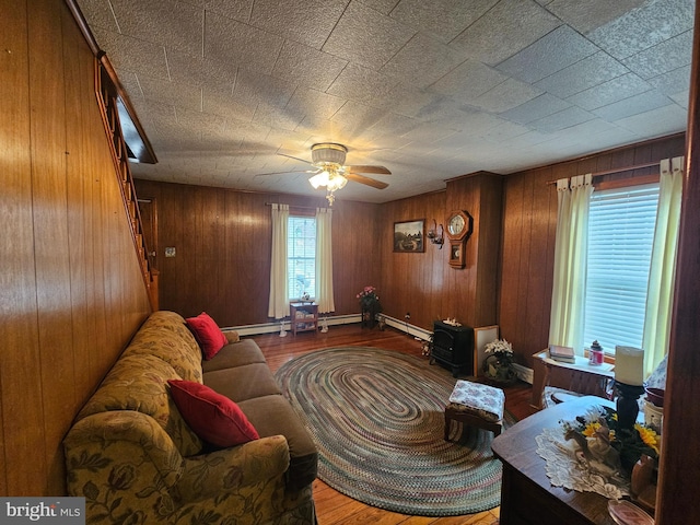 living area featuring ceiling fan, stairway, wood finished floors, wood walls, and a baseboard heating unit