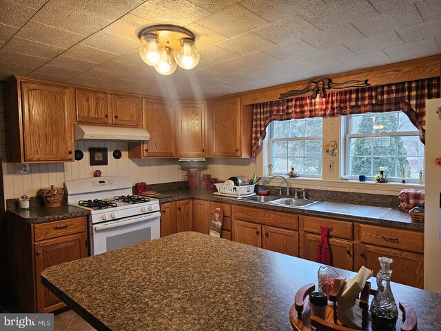 kitchen with under cabinet range hood, a sink, white gas range, brown cabinetry, and dark countertops