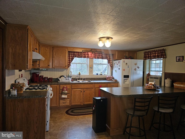 kitchen with under cabinet range hood, white appliances, a sink, brown cabinetry, and dark countertops