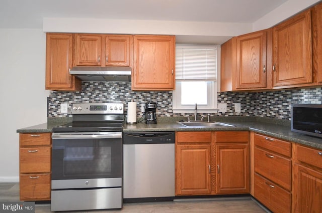 kitchen featuring brown cabinets, dark countertops, appliances with stainless steel finishes, a sink, and under cabinet range hood