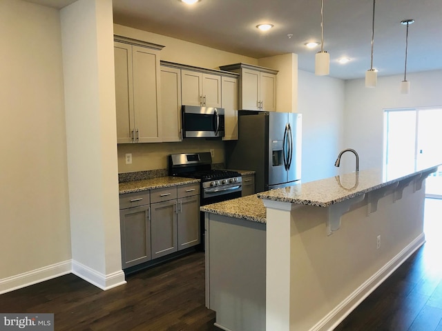 kitchen with gray cabinets, appliances with stainless steel finishes, hanging light fixtures, light stone countertops, and a center island with sink