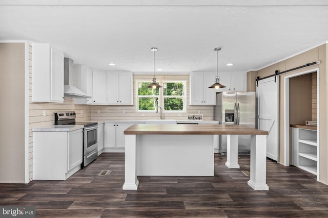 kitchen featuring wall chimney exhaust hood, decorative light fixtures, appliances with stainless steel finishes, a barn door, and white cabinets