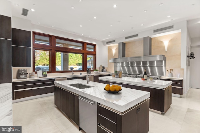 kitchen featuring sink, light stone counters, wall chimney range hood, a kitchen island with sink, and backsplash