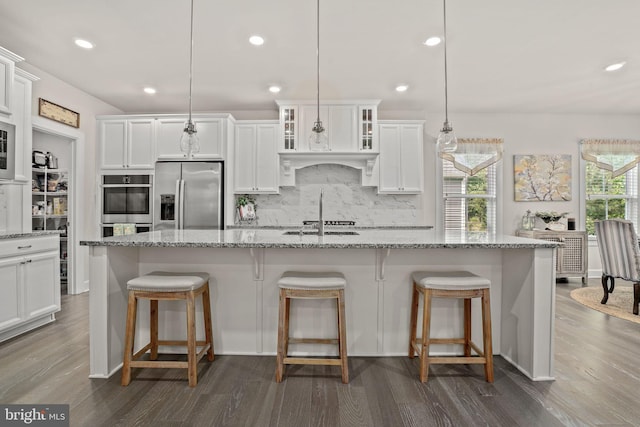 kitchen with white cabinets, hanging light fixtures, light stone counters, stainless steel appliances, and a center island with sink