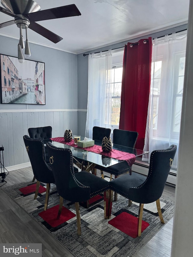 dining area featuring a wainscoted wall, crown molding, baseboard heating, ceiling fan, and wood finished floors