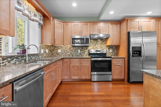 kitchen with sink, appliances with stainless steel finishes, hardwood / wood-style flooring, dark stone counters, and backsplash