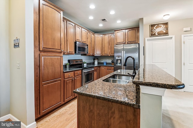 kitchen with sink, appliances with stainless steel finishes, dark stone countertops, light hardwood / wood-style floors, and a center island with sink