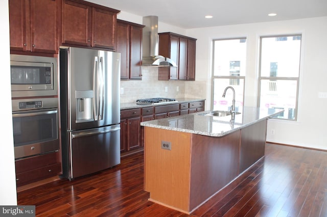 kitchen featuring wall chimney exhaust hood, sink, light stone counters, appliances with stainless steel finishes, and an island with sink