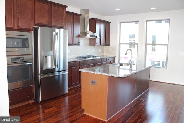 kitchen with wall chimney range hood, sink, appliances with stainless steel finishes, a kitchen island with sink, and light stone countertops