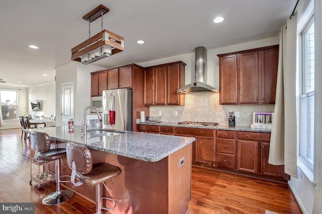 kitchen featuring stainless steel appliances, an island with sink, light stone counters, and wall chimney exhaust hood