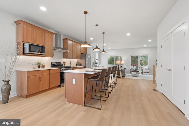 kitchen with hanging light fixtures, a kitchen island with sink, wall chimney exhaust hood, stainless steel appliances, and light hardwood / wood-style flooring