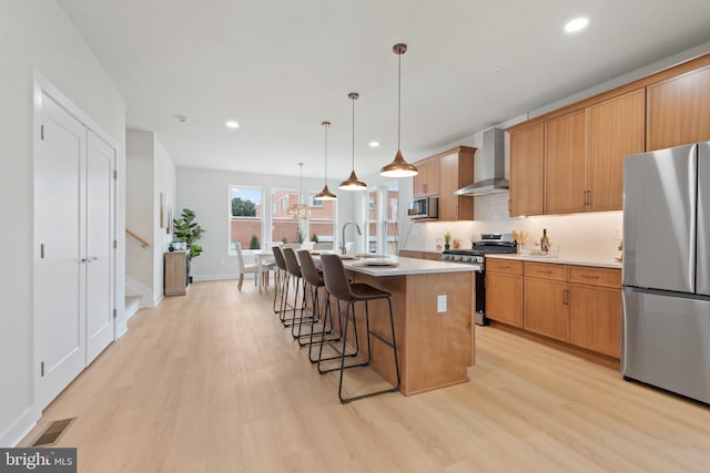 kitchen with light wood-type flooring, appliances with stainless steel finishes, an island with sink, pendant lighting, and wall chimney range hood