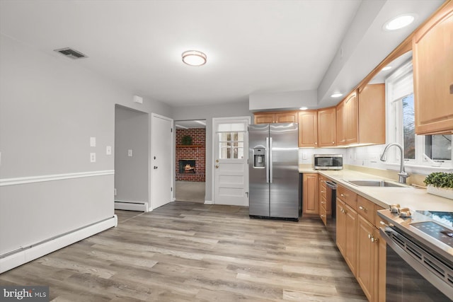 kitchen with sink, baseboard heating, appliances with stainless steel finishes, a fireplace, and light hardwood / wood-style floors