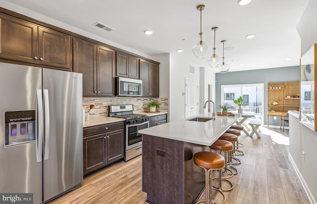 kitchen featuring sink, appliances with stainless steel finishes, a kitchen island with sink, hanging light fixtures, and dark brown cabinetry