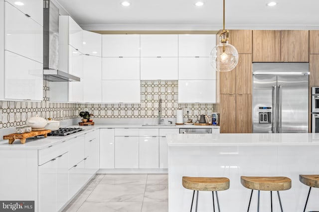 kitchen featuring a kitchen bar, white cabinetry, decorative light fixtures, stainless steel appliances, and wall chimney range hood