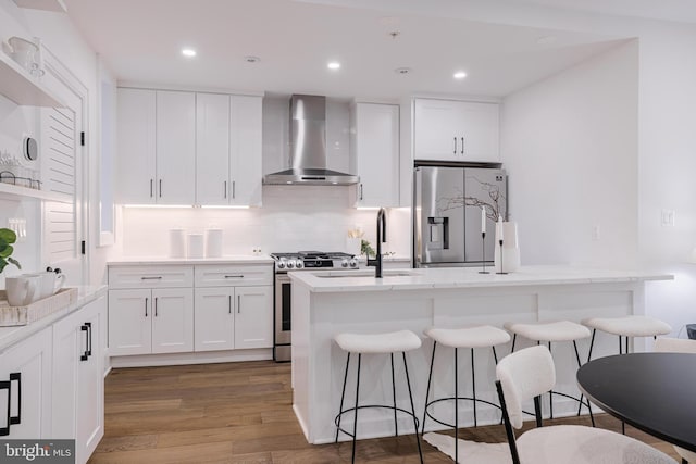 kitchen featuring wall chimney exhaust hood, a kitchen bar, a center island with sink, appliances with stainless steel finishes, and white cabinets