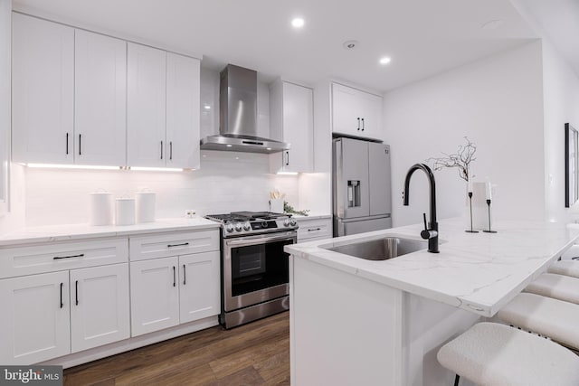 kitchen with sink, appliances with stainless steel finishes, a kitchen island with sink, white cabinetry, and wall chimney exhaust hood