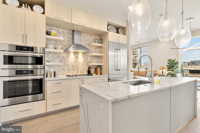 kitchen featuring decorative light fixtures, a center island with sink, stainless steel appliances, light stone countertops, and wall chimney range hood