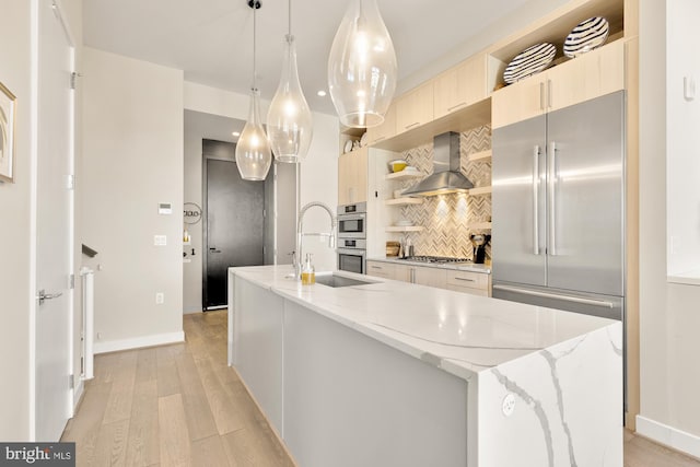 kitchen featuring wall chimney exhaust hood, light stone counters, an island with sink, pendant lighting, and stainless steel appliances