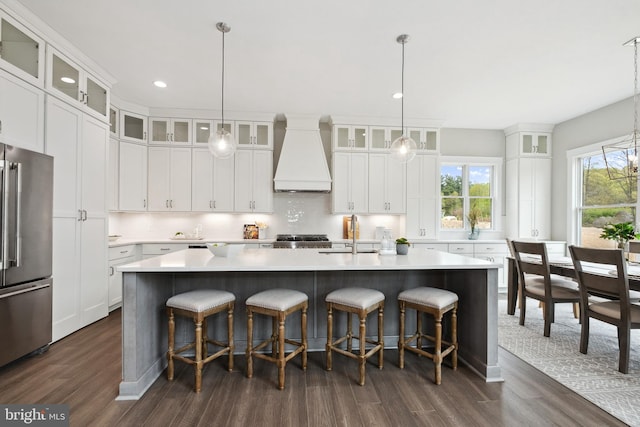 kitchen featuring sink, stainless steel refrigerator, custom range hood, an island with sink, and white cabinets