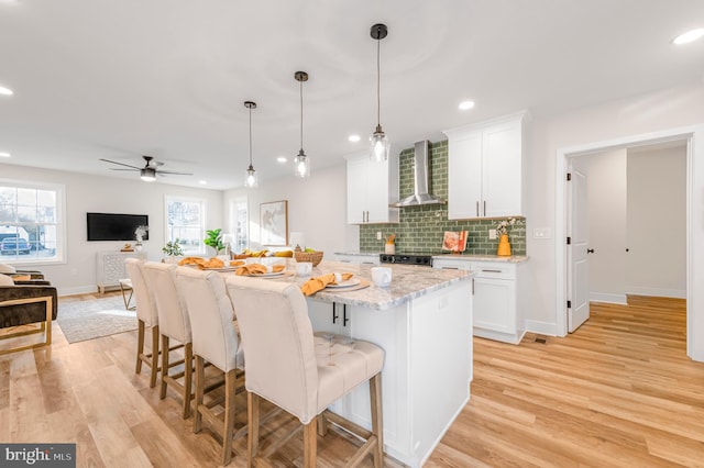 kitchen featuring wall chimney range hood, white cabinetry, hanging light fixtures, a center island, and light stone countertops