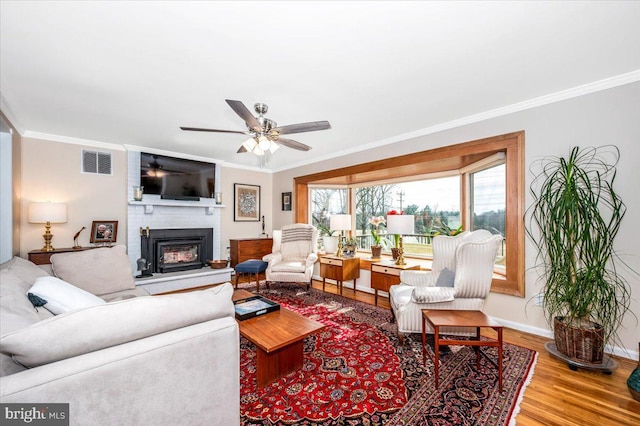 living area featuring wood finished floors, visible vents, baseboards, a brick fireplace, and crown molding