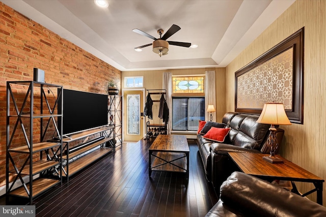 living room with dark wood-type flooring, a raised ceiling, ceiling fan, and brick wall