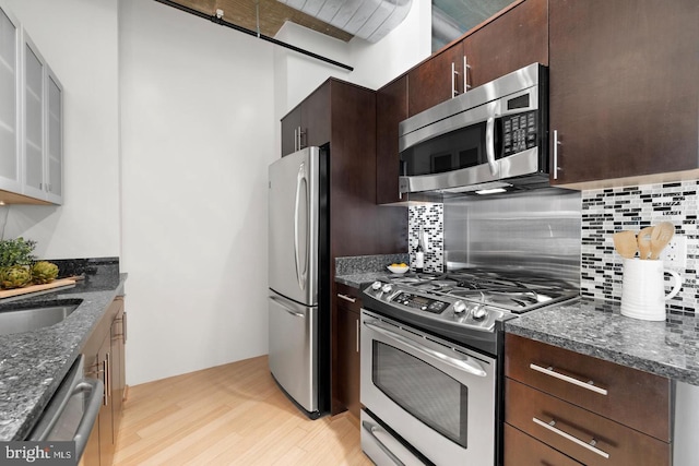 kitchen with dark stone countertops, dark brown cabinets, stainless steel appliances, tasteful backsplash, and light wood-type flooring