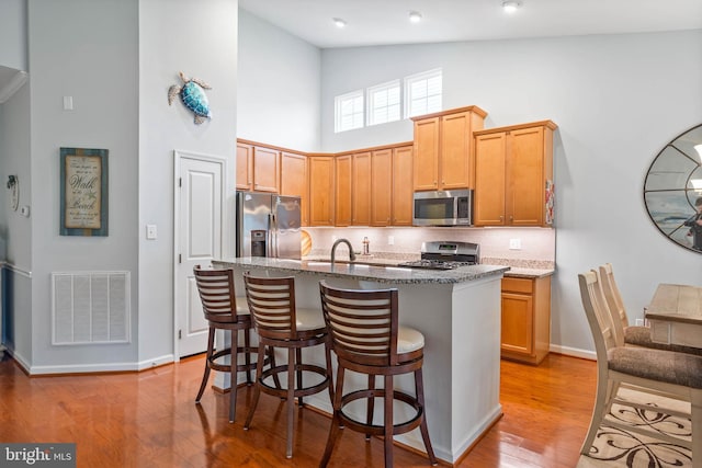 kitchen featuring sink, high vaulted ceiling, stainless steel appliances, light stone counters, and a center island with sink