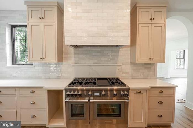 kitchen with range with two ovens, tasteful backsplash, cream cabinets, wall chimney exhaust hood, and light wood-type flooring