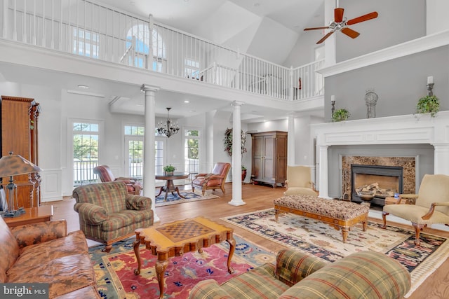 living room with ceiling fan with notable chandelier, light wood-style floors, a fireplace, decorative columns, and a towering ceiling