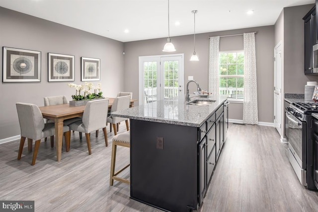 kitchen with sink, stainless steel gas range oven, light stone counters, decorative light fixtures, and a kitchen island with sink