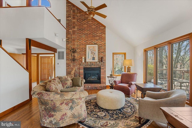 living area with a brick fireplace, high vaulted ceiling, stairs, a ceiling fan, and wood finished floors
