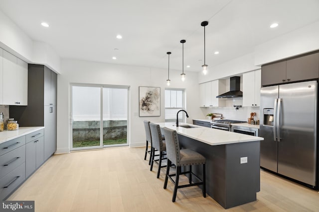 kitchen with appliances with stainless steel finishes, a kitchen island with sink, hanging light fixtures, white cabinets, and wall chimney exhaust hood