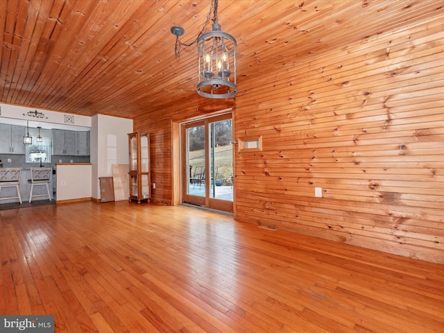 unfurnished living room with light wood-type flooring, an inviting chandelier, wooden ceiling, and wood walls