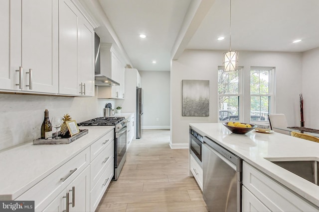 kitchen featuring appliances with stainless steel finishes, decorative light fixtures, white cabinetry, wall chimney range hood, and light hardwood / wood-style flooring