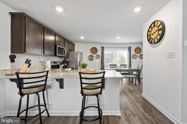 kitchen with a breakfast bar area, dark hardwood / wood-style flooring, dark brown cabinetry, stainless steel appliances, and light stone countertops