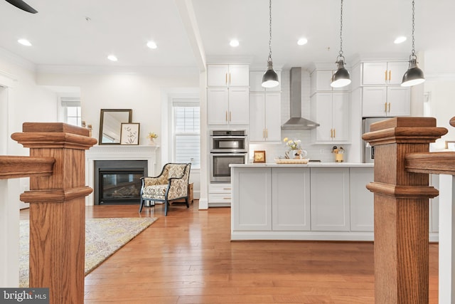 kitchen with tasteful backsplash, double oven, a glass covered fireplace, light wood-type flooring, and wall chimney exhaust hood