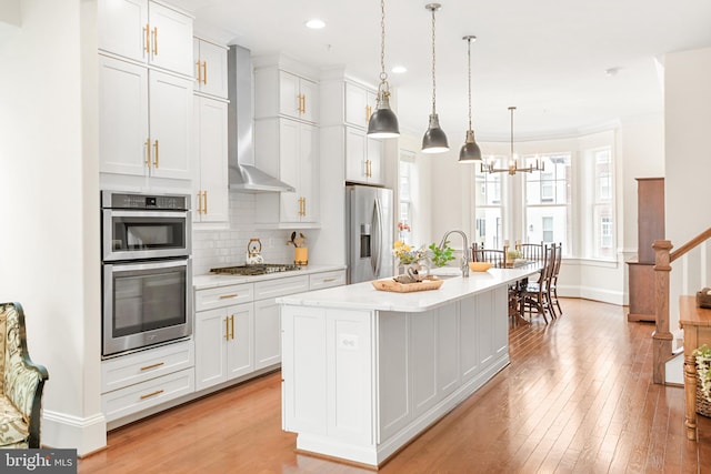 kitchen with a kitchen island with sink, stainless steel appliances, light countertops, wall chimney range hood, and backsplash