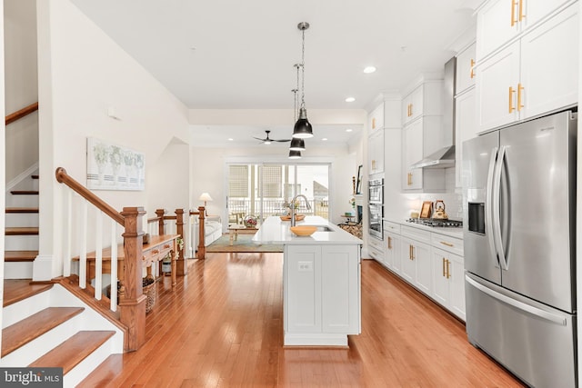 kitchen with appliances with stainless steel finishes, white cabinetry, a sink, wall chimney range hood, and light wood-type flooring